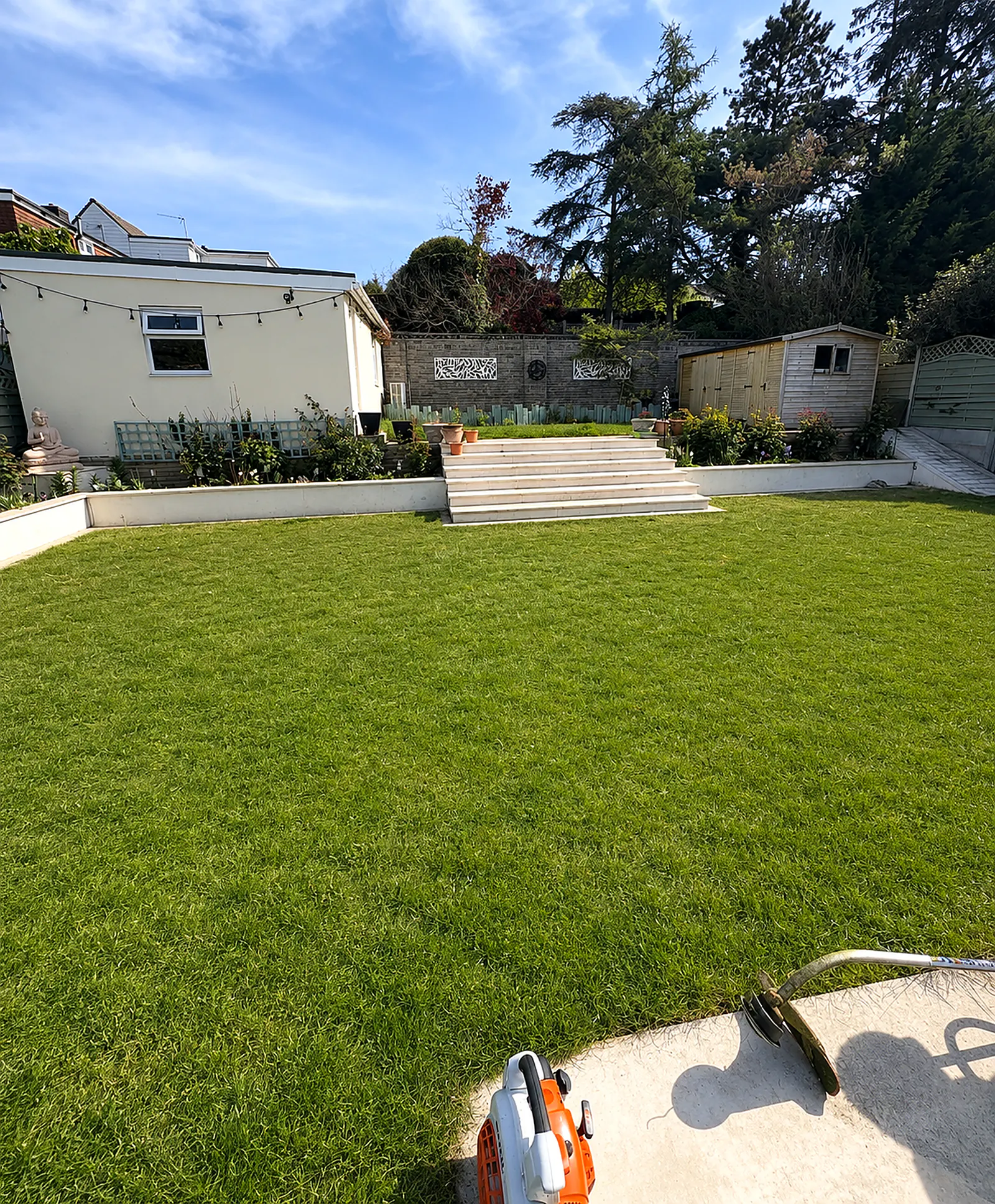 A modern stepped garden with a manicured lawn beneath a white render house with festoon lights